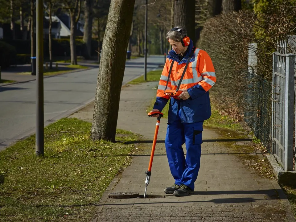 Técnico con audífonos caminando sobre una vereda con un micrófono de suelo para escuchar una fuga de agua