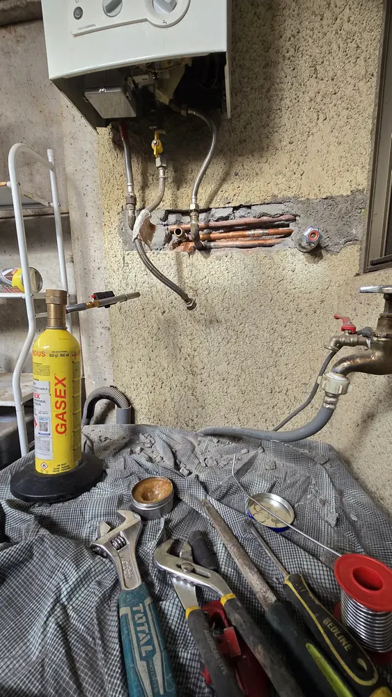 Cañerías de cobre a la vista en un muro bajo un calefont, con un cartucho de gas para soplete y herramientas
